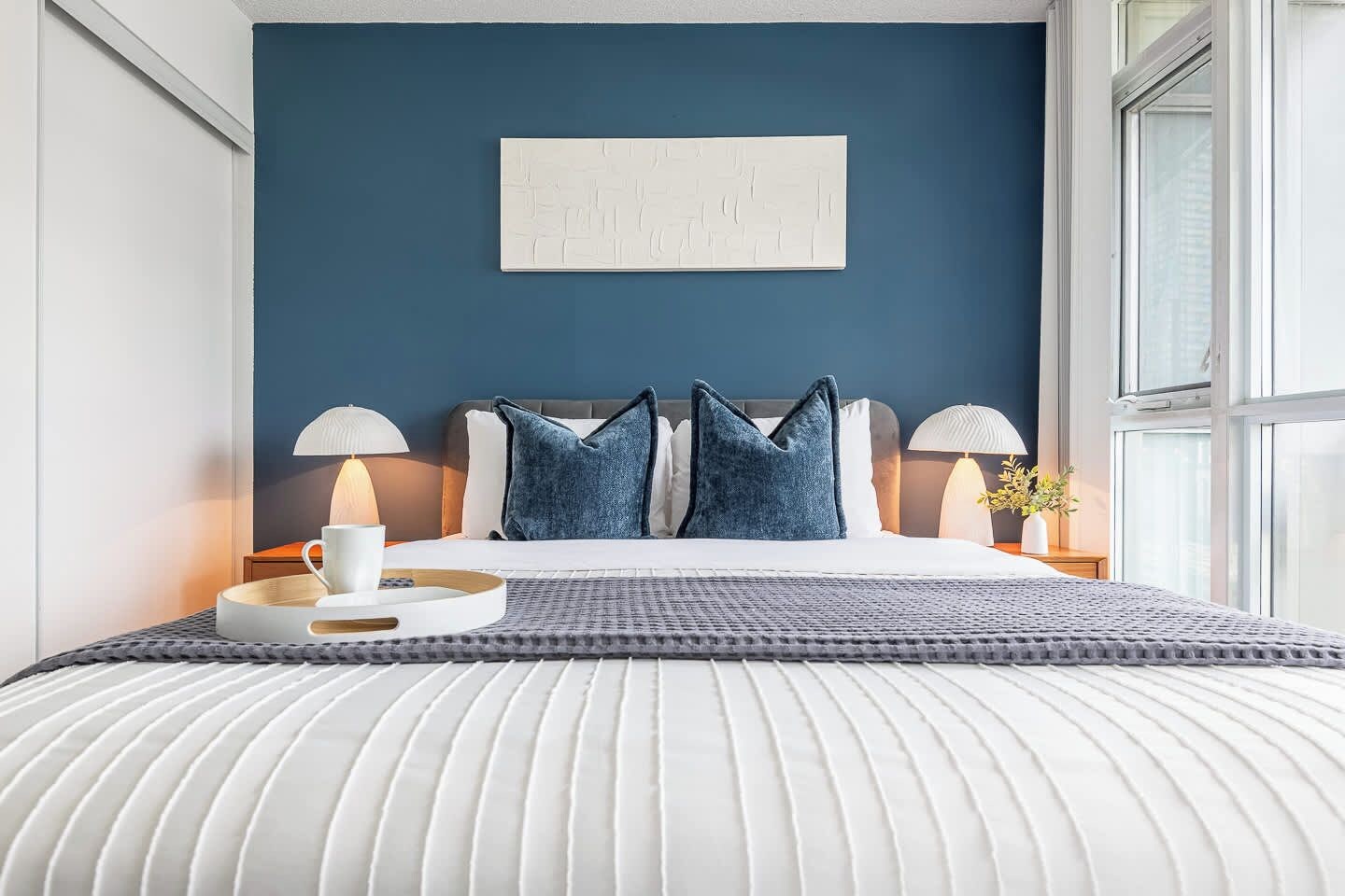 Een net opgemaakt bed met een wit hoofdbord, twee kussens en een blauw sierkussen. Er staat een nachtkastje met een lamp en een kleine vaas met bloemen.