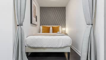 Quarto de hotel estreito com uma cama, mesa de cabeceira e uma luminária.