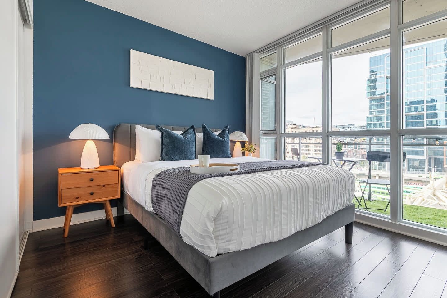 A modern bedroom with a large bed, a nightstand, a bedside table, and a view of the city through large windows.