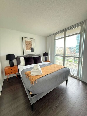 A modern hotel room with a large bed, two bedside tables, a city view, and a framed artwork on the wall.