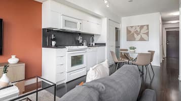 Uma sala de estar moderna com um sofá, uma mesa de centro de vidro e uma área de cozinha com armários brancos e um fogão.