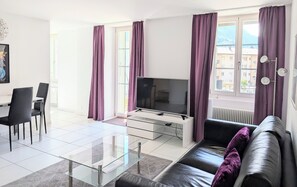 A modern living room with a glass coffee table, a black leather sofa, a flat-screen TV, and a dining area with chairs. - The Centralstrasse Apartment | Heart of Interlaken (Interlaken)