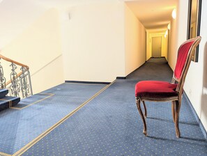 A hallway with a blue carpet, a red upholstered chair, and a staircase with ornate metal railings. - The Centralstrasse Apartment | Heart of Interlaken (Interlaken)