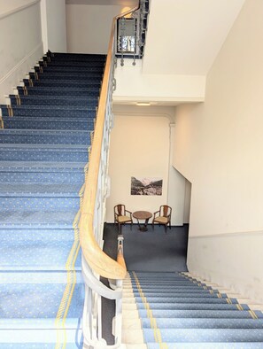 A staircase with blue carpeted steps and a handrail, leading to a room with two chairs and a table. - The Centralstrasse Apartment | Heart of Interlaken (Interlaken)