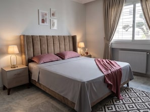 Ein Schlafzimmer mit einem Bett, Nachttischen, Lampen, einem Heizkörper und einem Fenster mit Vorhängen.
