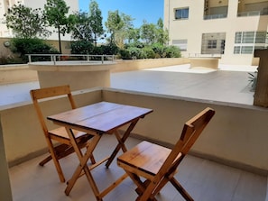 Ein Patio im Freien mit einem Holztisch und -stühlen, einem weiß gefliesten Boden und Blick auf ein Gebäude und Bäume im Hintergrund.