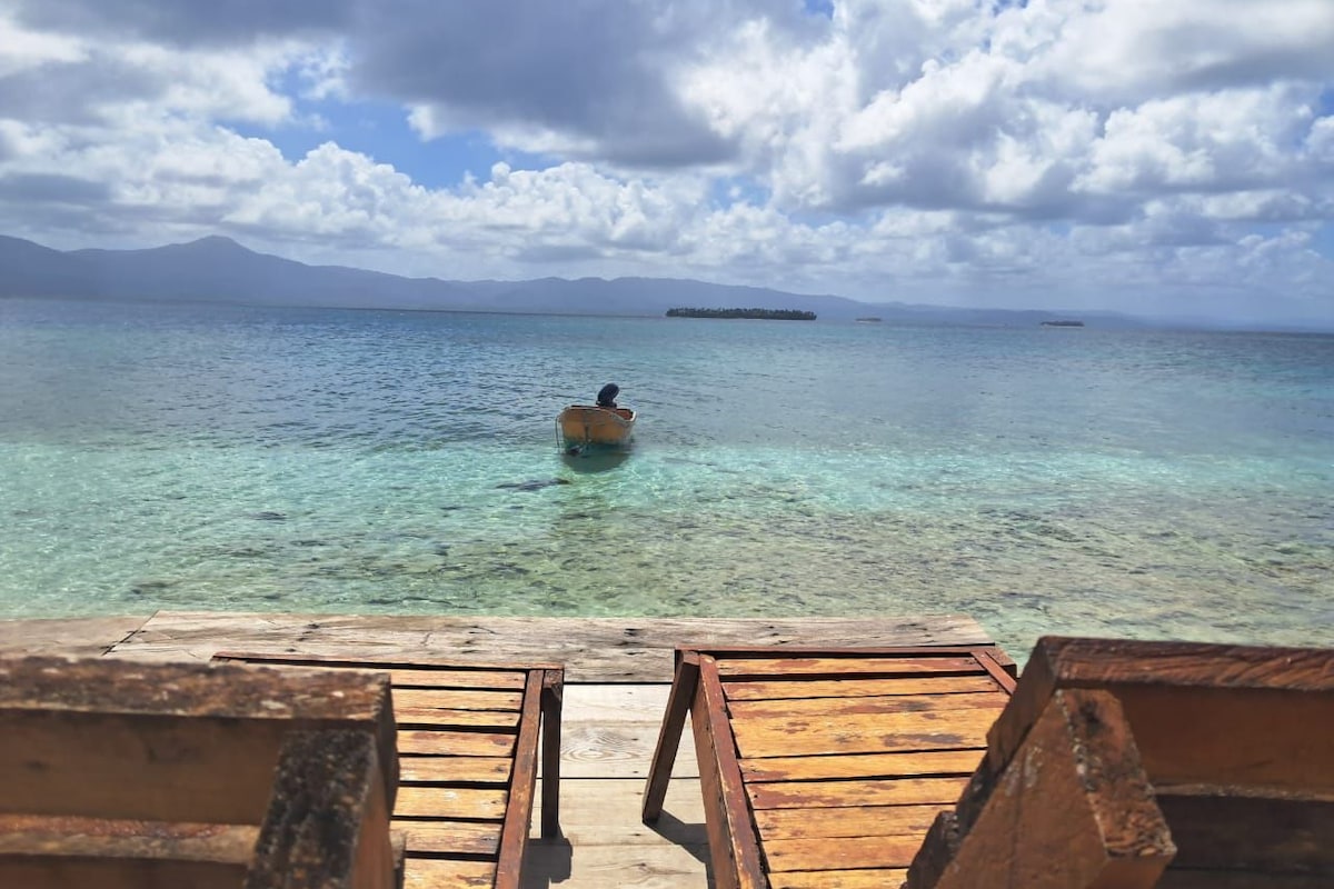 Una terraza de madera con vista a un mar azul y despejado, con una persona en un bote a lo lejos.