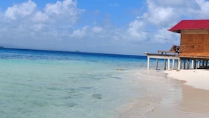 A beach with clear turquoise water, a wooden structure with a red roof, and a white sandy shore. - Pelicano Island (Moraquedub)