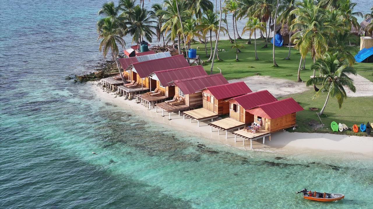 Una hilera de bungalows de madera con techos rojos en una playa, rodeados de palmeras y agua turquesa cristalina.