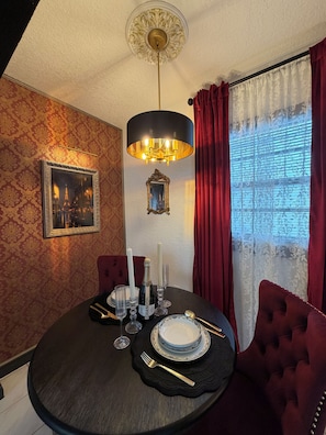 Een eethoek met een ronde tafel gedekt voor twee personen, met een zwarte tafelkleed, witte borden en bestek. Daarboven hangt een kroonluchter en aan de muur hangt een lijst met een afbeelding.