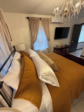 A hotel room with a bed, curtains, a chandelier, an air conditioning unit, a television, and a desk with a lamp.