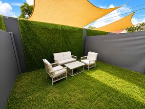 Eine Terrassenfläche mit einer weißen Outdoor-Sofagarnitur, einem kleinen Tisch und einem Sessel, umgeben von grünem Kunstgrün und einer Heckenwand.