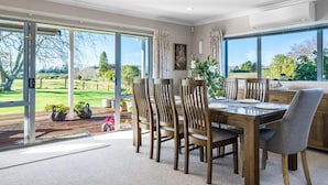A dining area with a table and chairs, large windows with a view of a green landscape, and a door leading outside. - The Pond House by Homello (Karaka)