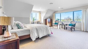 A bedroom with a bed, a nightstand, a chair, a small table, and a window with a view. - The Pond House by Homello (Karaka)