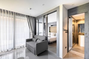 A modern hotel room with a grey sofa, a bed with a patterned headboard, and a bathroom with a mirror and sink. - Kamala - Peaceful Courtyard (Kathu District)