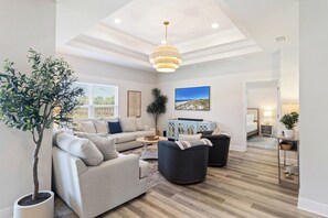 A modern living room with a beige sofa, a dark armchair, and a coffee table. There is a large window with blinds, a potted plant, and a painting on the wall. - Luxurious-GolfCart-OceanView-BeachGear-Games (Port St. Joe)