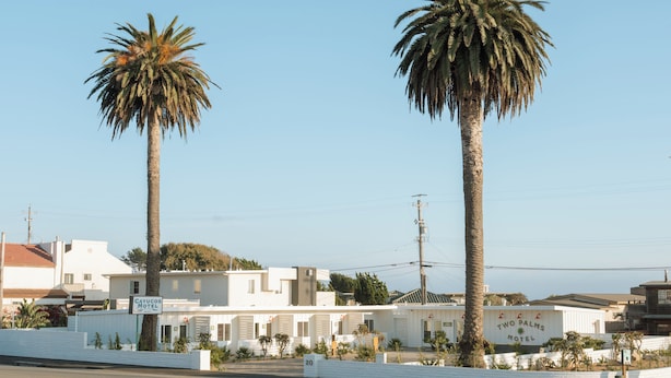 A motel with two prominent palm trees, a white building with a sign, and a clear sky. - Two Palms Motel (Cayucos)