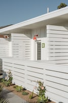 Un edificio blanco con listones horizontales, una puerta con un letrero de número y un ventanal pequeño.