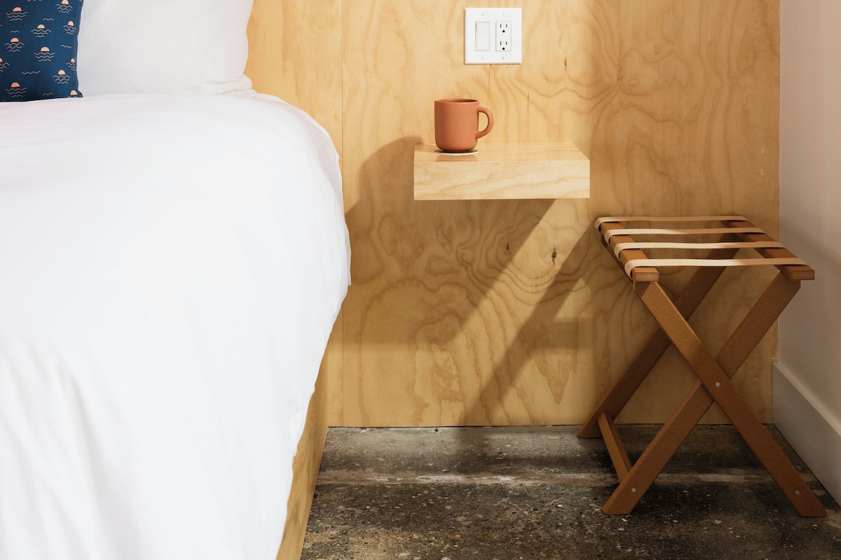 A bedroom with a patterned wallpaper, a bedside table with a mug, a green lamp, and a wooden shelf.