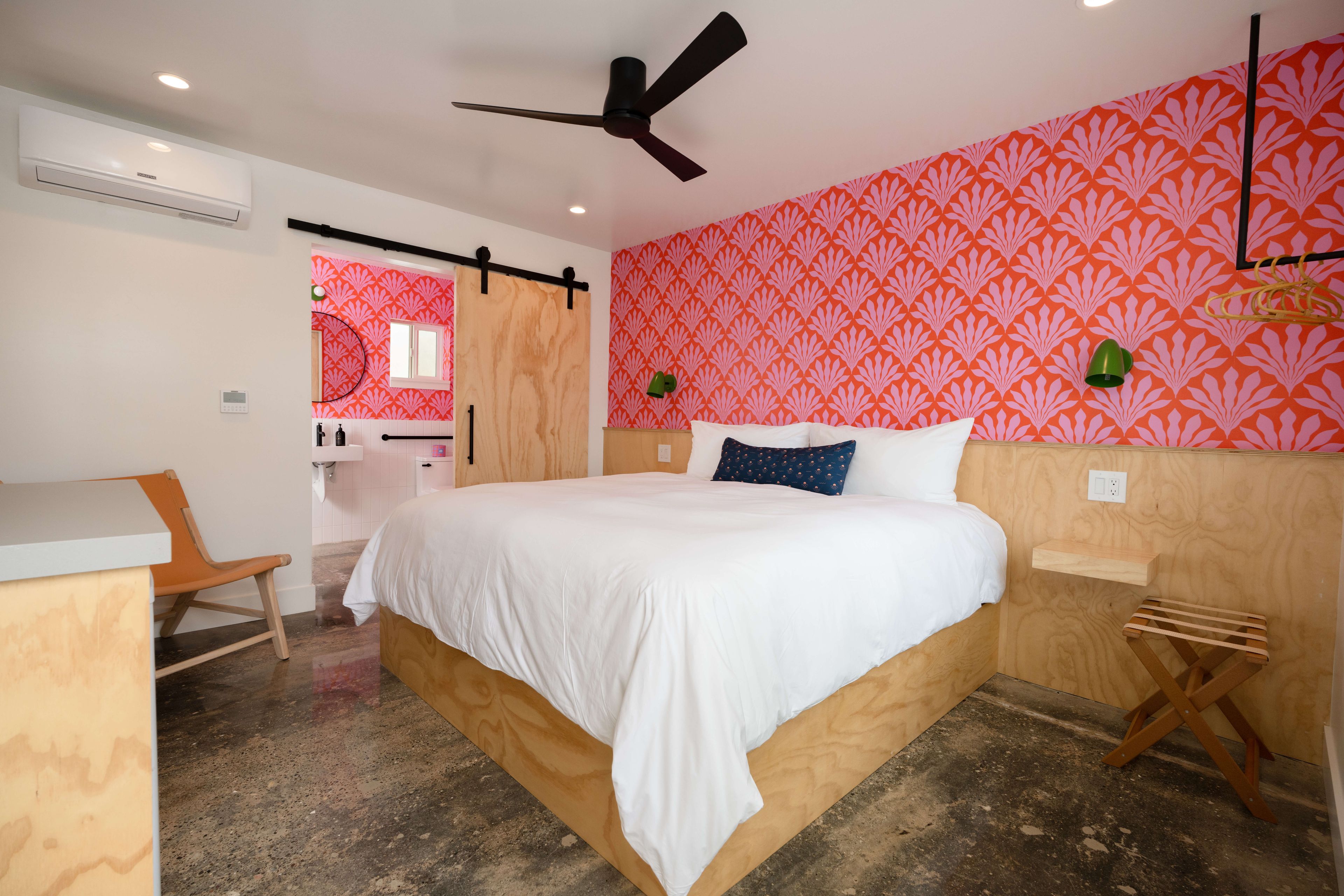 A bedroom with a bed, a desk, a chair, a ceiling fan, and a wall with a patterned wallpaper.