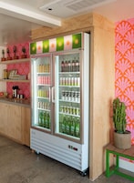 A beverage refrigerator with multiple shelves stocked with drinks, a wooden counter with a coffee machine, and a potted plant on a table.
