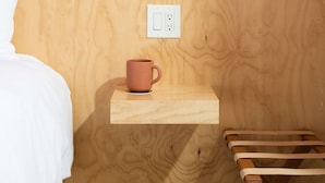 A bedroom with a patterned wallpaper, a bedside table with a mug, a lamp, and a wooden bench. - Two Palms Motel (Cayucos)