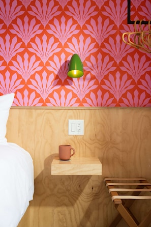 Quarto com papel de parede estampado, mesa de cabeceira com uma caneca, luminária e um banco de madeira.