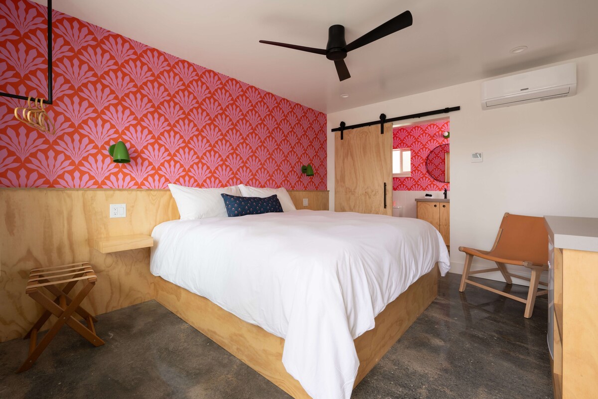 Quarto com cama, escrivaninha, cadeira, ventoinha no teto e banheiro visível através de uma porta deslizante.
