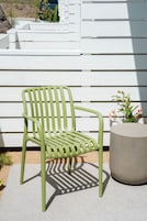 A green metal chair with a backrest and armrests, positioned on a concrete surface next to a white wall with horizontal siding.