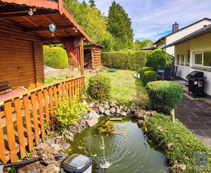 A wooden deck with a small pond, a barbecue area, and a well-maintained garden with hedges and trees.