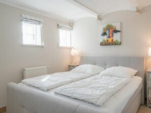A bedroom with a double bed, two windows with curtains, a radiator, a bedside table with a lamp, and a wall painting. - Kohstall in the Haubarg Ossenhoff (Tating)