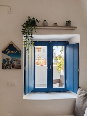 A room with a blue door, a window seat, a wall-mounted shelf with plants, and a framed picture of Greece. - Casa Calma House (Emporio)