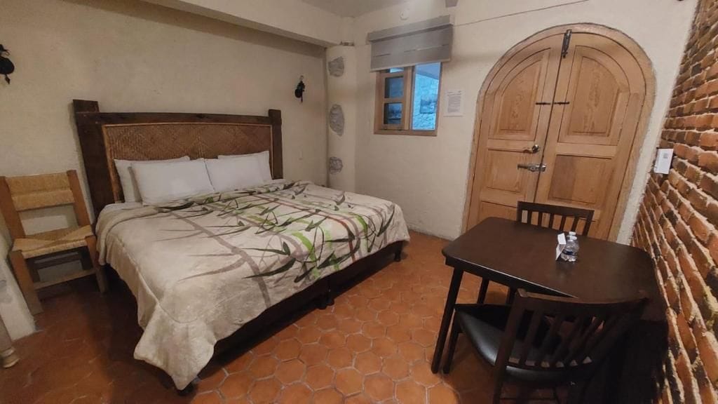 A bedroom with a bed, a wooden table with chairs, a small window, and a door.