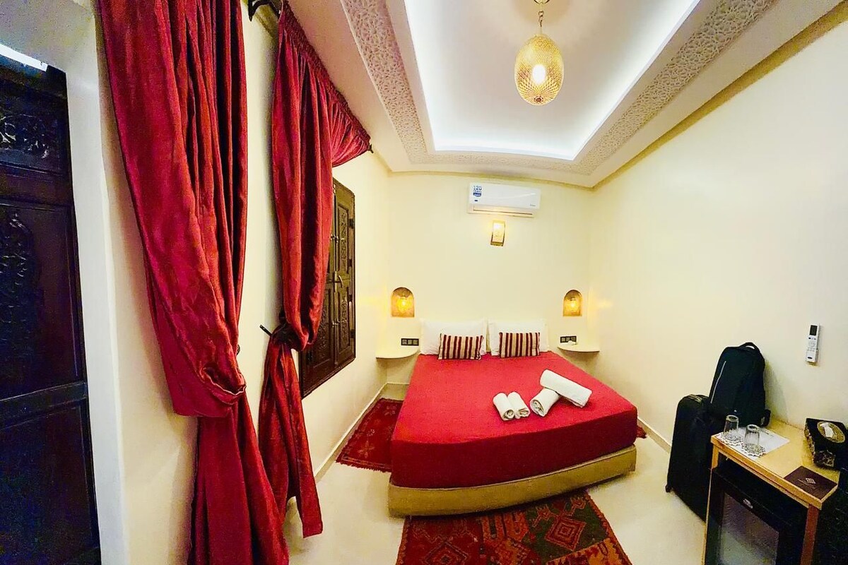A hotel room with a bed, curtains, a desk, a chair, a small table, and a wall-mounted air conditioner.