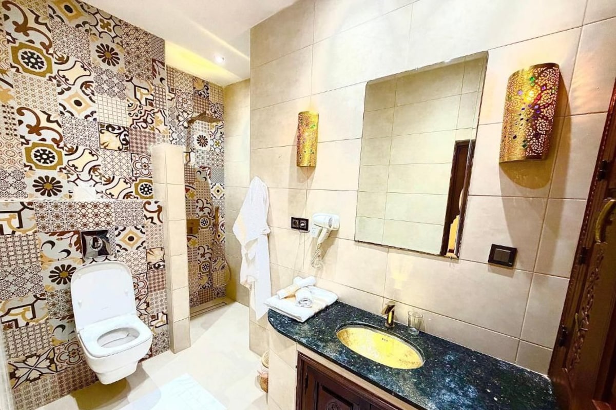 A bathroom with a patterned tile wall, a white toilet, a sink with a gold faucet, and a mirror with decorative lights.
