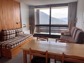 A room with a wooden floor, a sofa with plaid cushions, a wooden table, and a view of mountains through large windows. - Peisey-Loue: Apartment at the base of the slopes, sleeps 6 (Peisey-Nancroix)