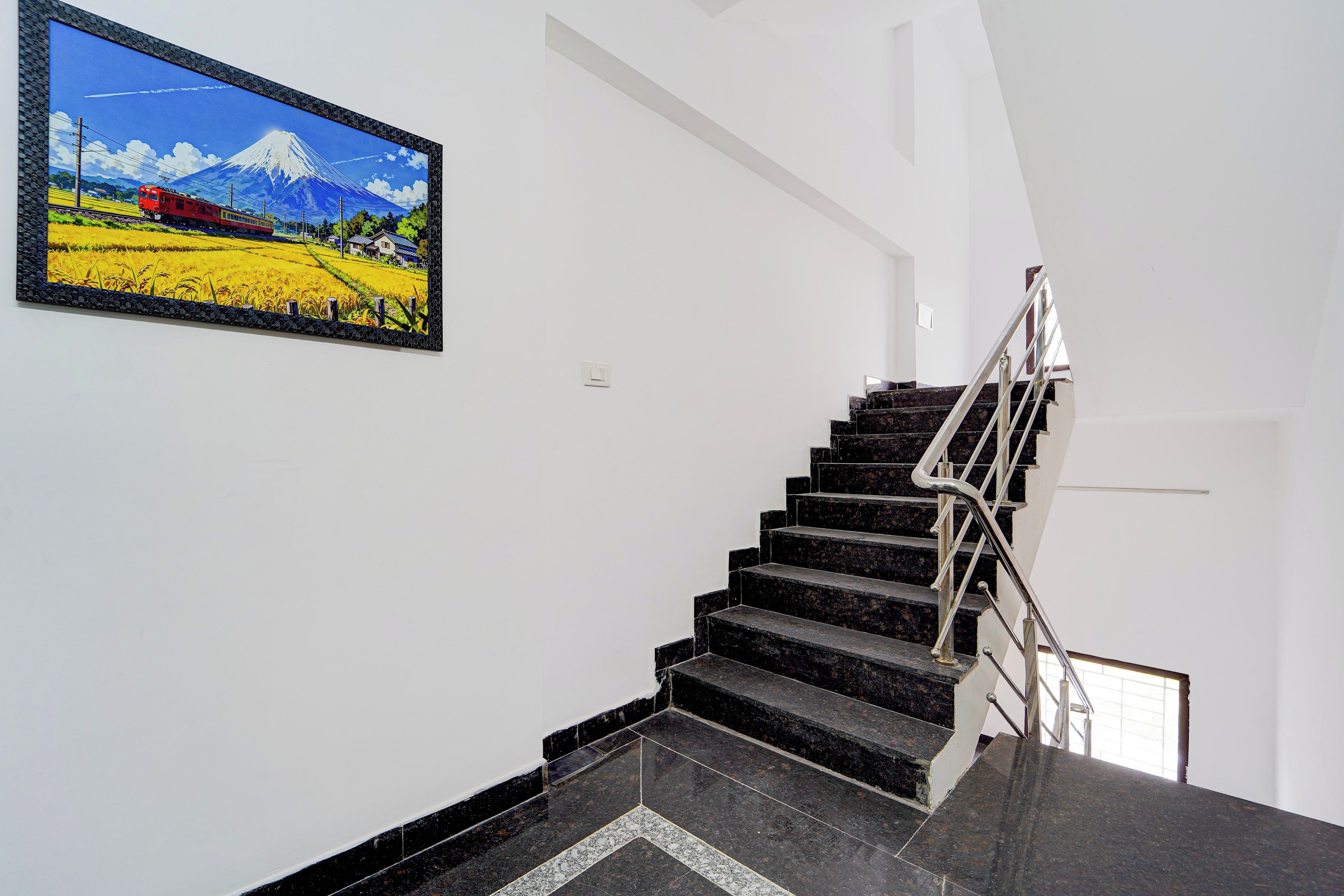 Una escalera con escalones negros y barandilla metálica, que lleva a un rellano con un cuadro enmarco de un paisaje.