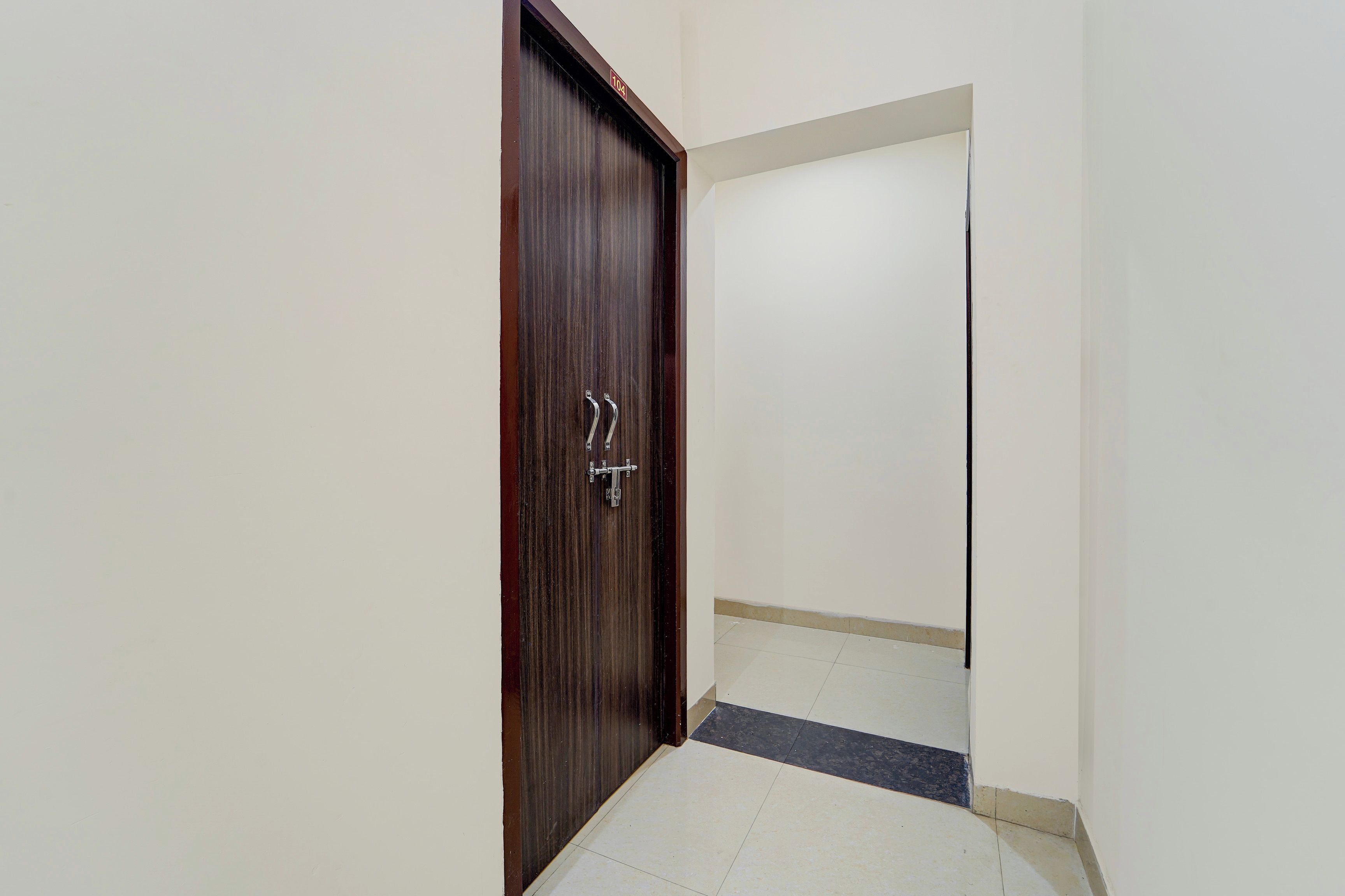 Un pasillo con una puerta de madera, piso de baldosas y pared blanca.