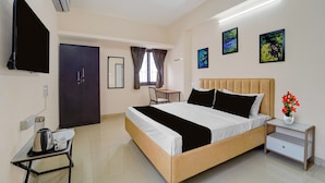 Quarto de hotel com cama, mesa de cabeceira, escrivaninha com cadeira, televisão e obras de arte nas paredes.