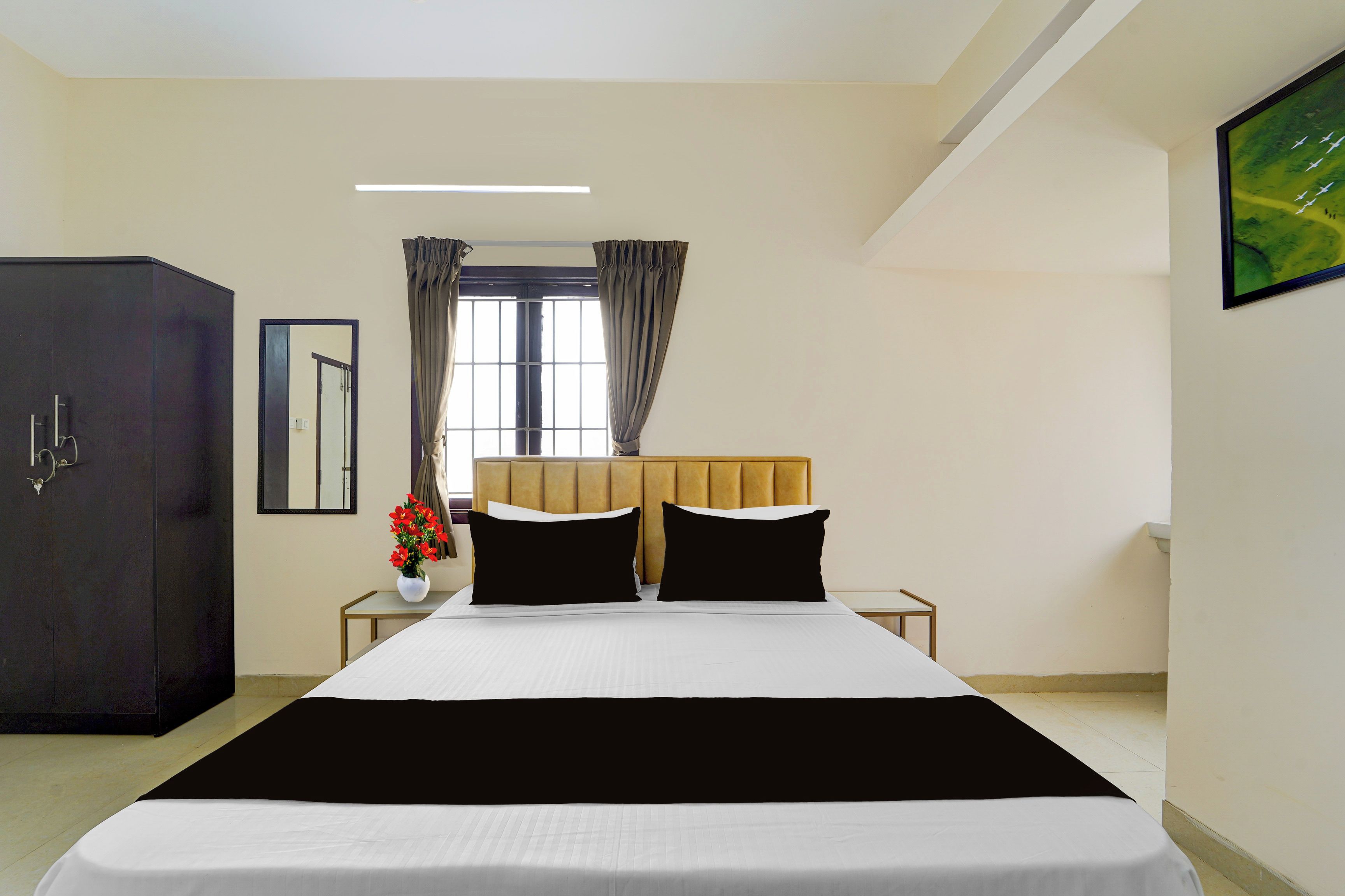 A hotel room with a bed, a wardrobe, a nightstand with a vase of flowers, and a window with curtains.