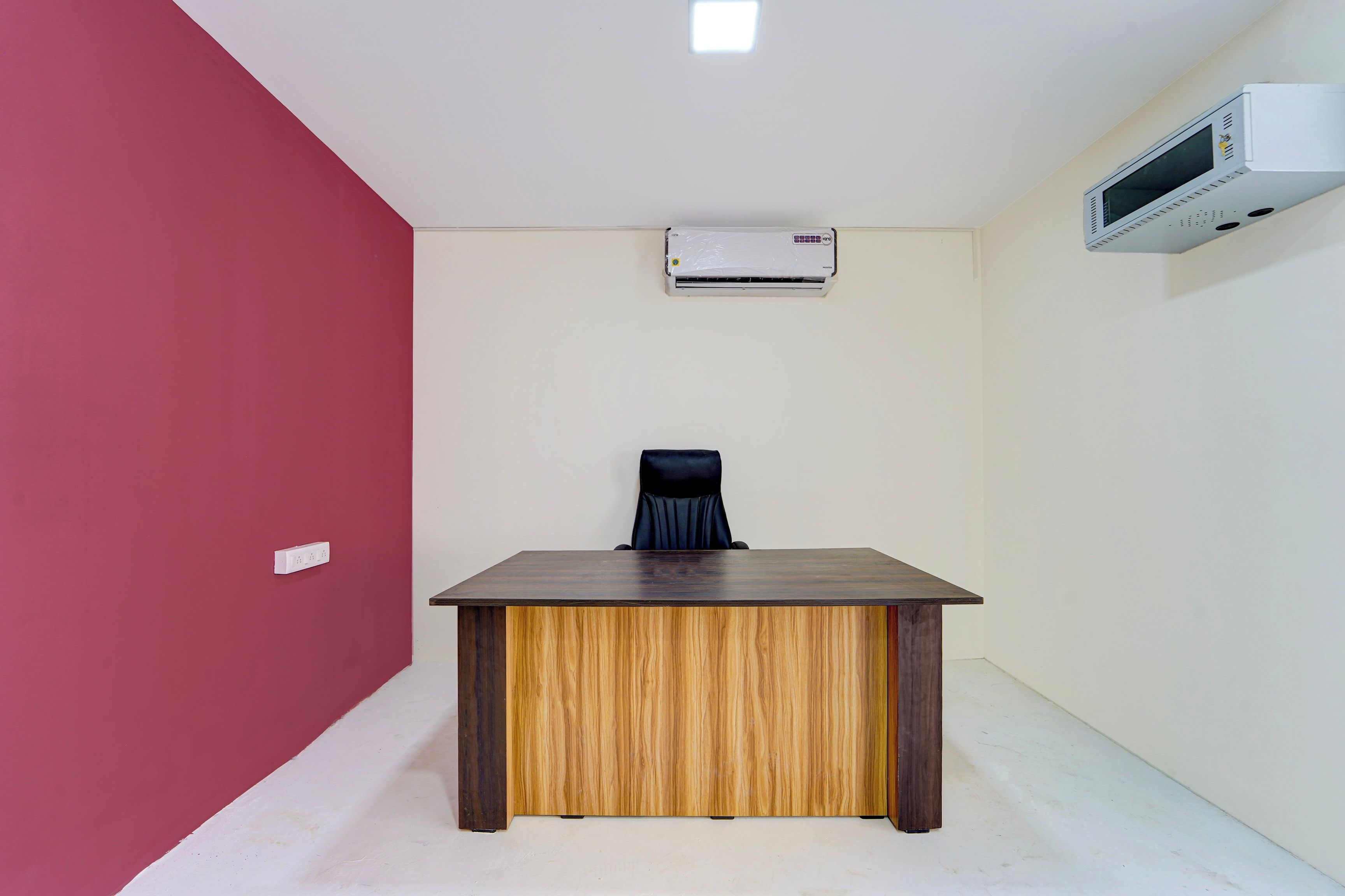 A room with a wooden desk, a black office chair, a white air conditioning unit, and a white wall.