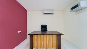 A room with a wooden desk, a black office chair, a white air conditioning unit, and a white wall.