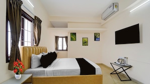 Quarto de hotel com cama, mesa de cabeceira, uma pequena mesa com chaleira, televisão de tela plana e uma obra de arte na parede.
