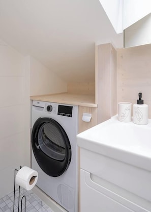 A compact laundry room with a front-loading washing machine, a toilet paper holder, and a small shelf with two toiletry bottles. - Stylish 2-BR Loft | AC, Cheap parking & Sleeps 6 (Riga)