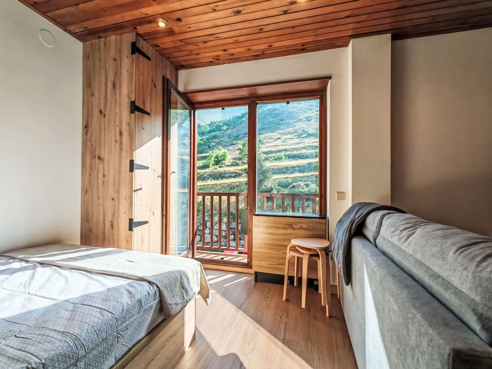 A bedroom with a wooden ceiling, a bed, a sofa, and a balcony with a view of a hilly landscape.