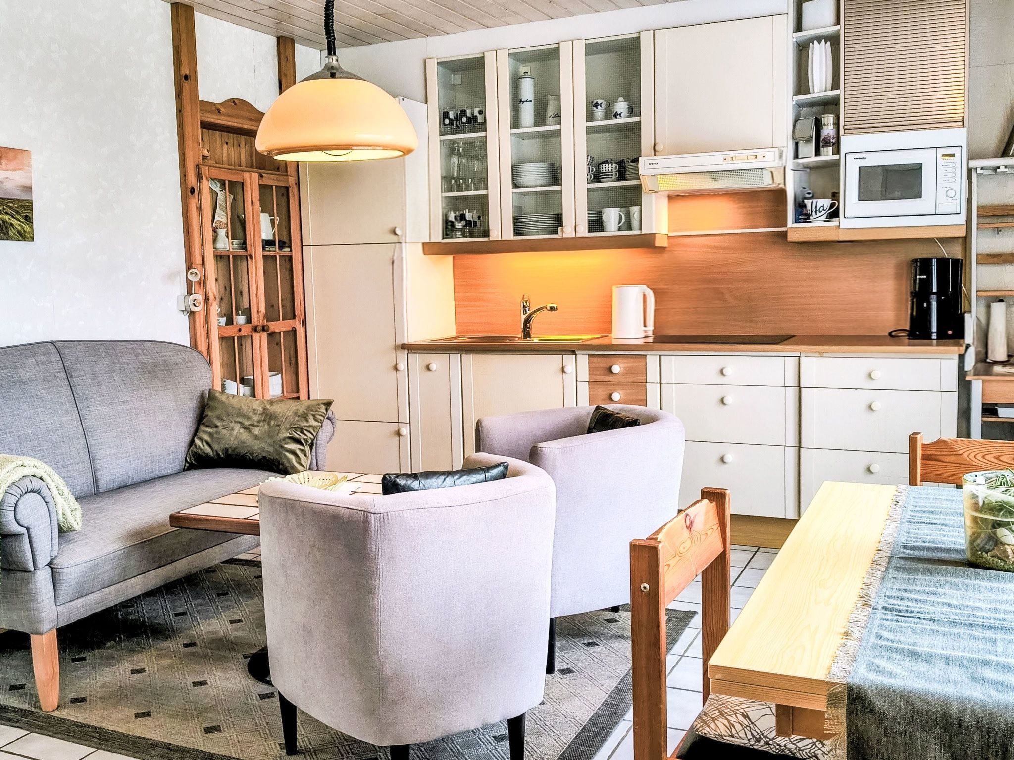 Una cucina moderna con mobili bianchi, un forno a microonde e una macchina da caffè. Nella zona giorno, un divano grigio e due poltrone.