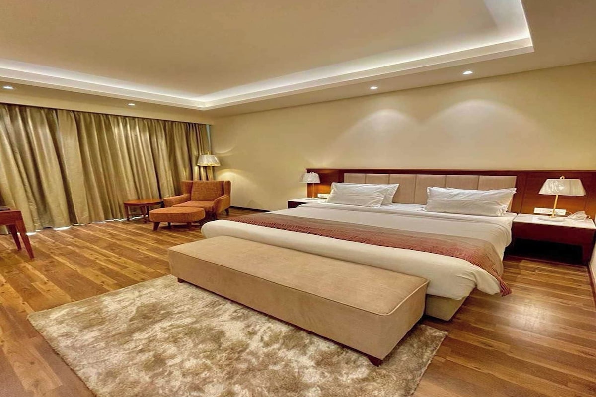 Une chambre d’hôtel avec un grand lit, un coin salon comprenant une chaise et une table, un banc et une grande fenêtre avec des rideaux.