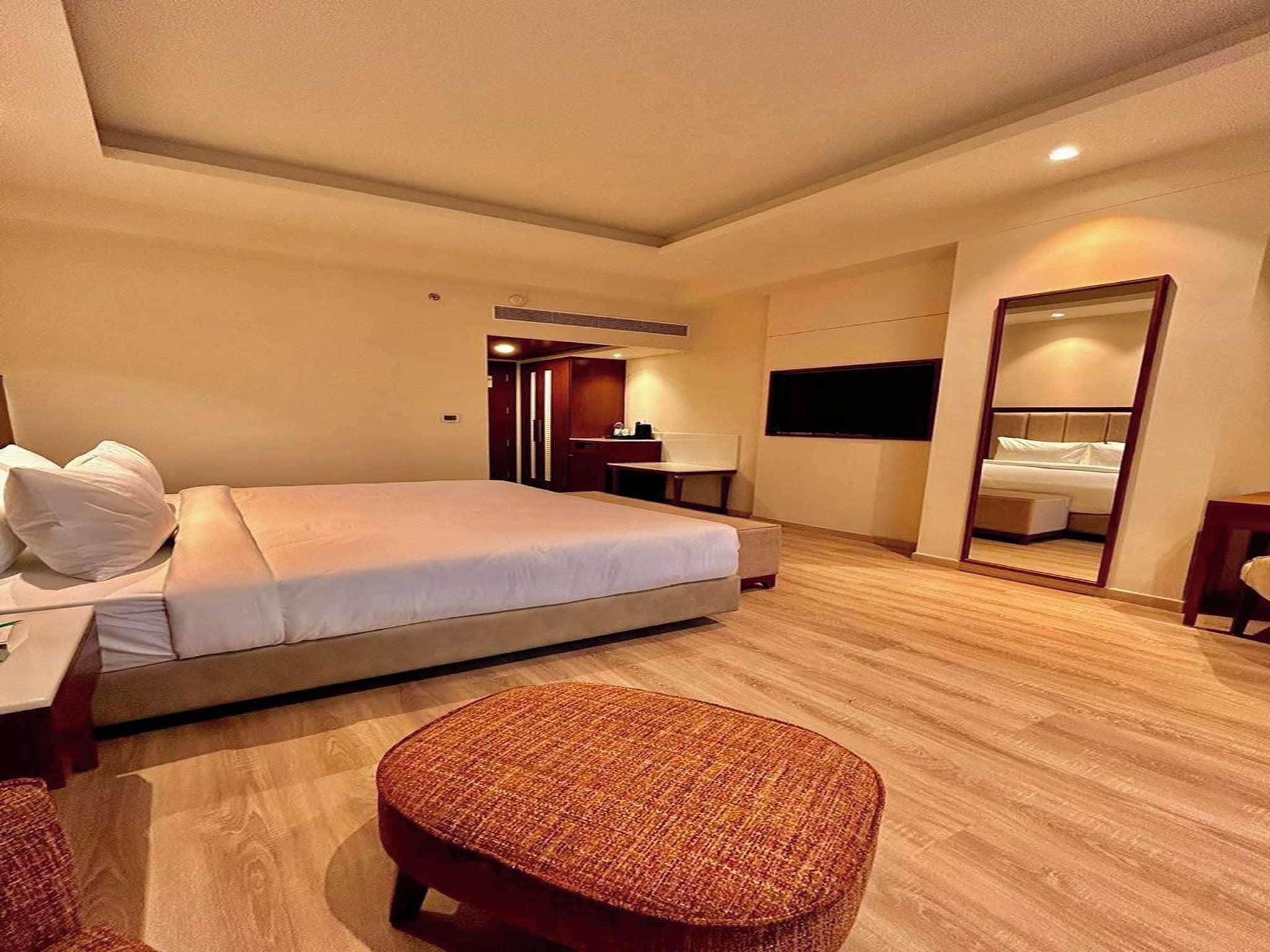 A hotel room with a bed, a desk, a chair, a mirror, and a small ottoman.