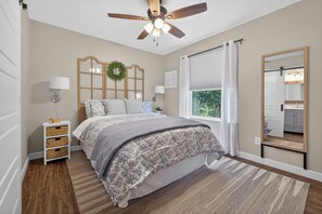 Ein Schlafzimmer mit Bett, Nachttisch, Wandlampe, Deckenventilator, Fenster mit Vorhängen und einem Spiegel.