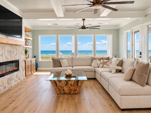 A living room with a stone fireplace, a glass coffee table, a beige sofa, and a view of the ocean through large windows.